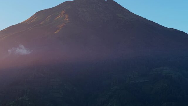 Aerial view of the majestic Mount Sumbing peak during sunrise with mist and golden light over the slopes near Kecamatan Kalikajar, Jawa Tengah, Indonesia.