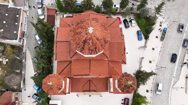High-angle aerial tracking shot moving forward directly over the textured red-tiled roofs, central cross dome, and twin bell towers of the modern Orthodox Cathedral in Berat, Albania