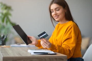 Fototapeta premium Smiling Young Woman Holding Credit Card And Tablet, Enjoying Online Shopping From Comfortable Home Office. Engaging In Digital Shopping Experience While Seated At Modern Desk In Cozy Environment.