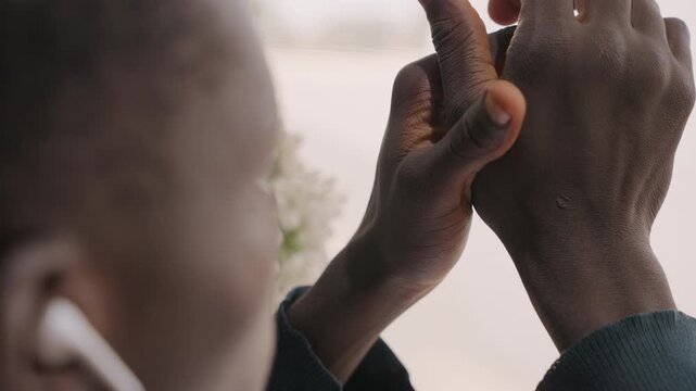 Close up black man listening earbuds, fingers fidgeting with hands, floral window background, soft daylight, private connection, checking messages, rehearsing voice, affectionate memory, calm
