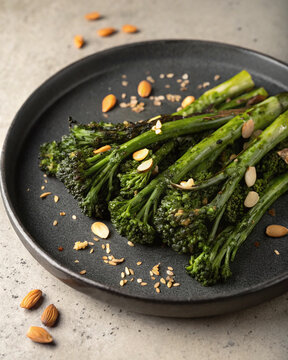 Photorealistic charred broccolini on dark ceramic plate with garlic confit lemon zest almonds rustic grill lighting composition