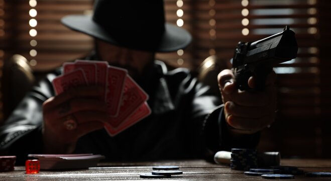 Mafia. Man with gun and playing cards at wooden table in dark room, selective focus