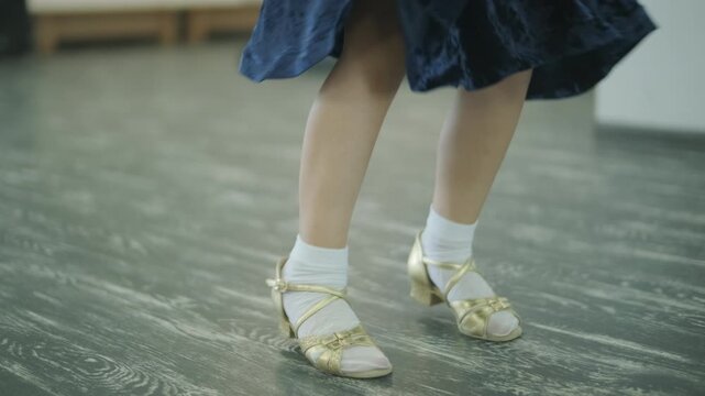 Child dancer's feet in golden high heel shoes training on a parquet floor during choreography lesson.