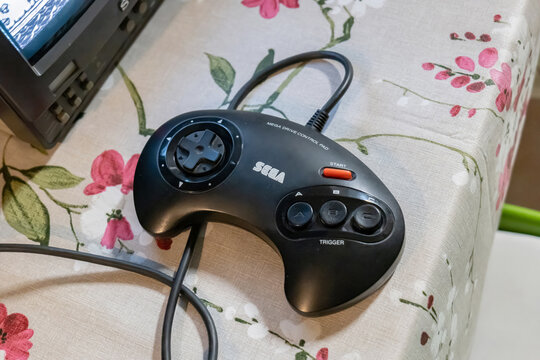Black Sega Mega Drive control pad on floral tablecloth beside an old CRT television, wired retro game controller close-up for classic 1990s console gaming nostalgia, no people