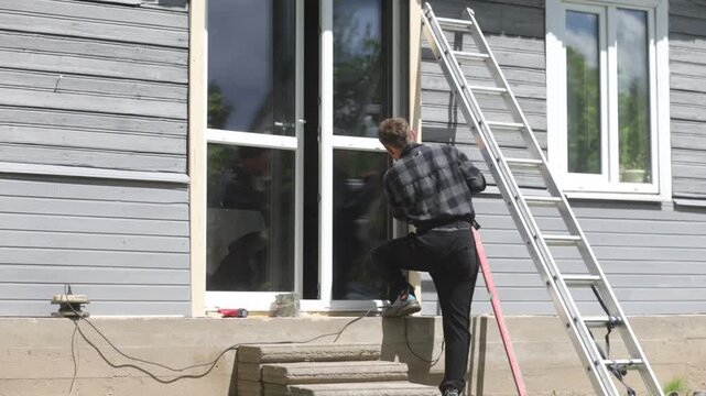 A man is renovating a wooden house, drilling a hole in the frame of a glass door outside the house