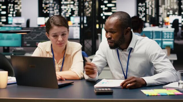Corporate staff collaborating in a shared workspace late at night, engaged in teamwork and planning sessions. Multiethnic group uses laptop to analyze data and discuss strategies, action plan.