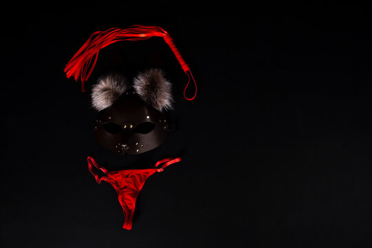Red lace thong panties, a brown leather cat mask with fluffy faux fur pom-poms, and a red leather flogger whip isolated on a black textured surface with empty space.