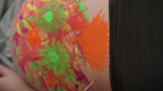 Closeup pregnant white woman belly detail painted, macro view of neon green and orange paint texture, brushstroke movement and glitter beads catching light, hand steadying palette, studio vibe,
