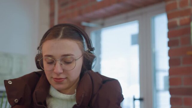 White woman in cozy cafe, wearing headphones and round glasses, bundled in brown puffer coat, seated by large window with exposed brick, listening intently with relaxed contemplative expression, soft