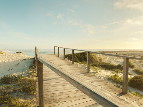 Serene wooden boardwalk leading through sunlit coastal dunes to the vast Atlantic Ocean at Furadouro Beach during a tranquil golden hour sunset