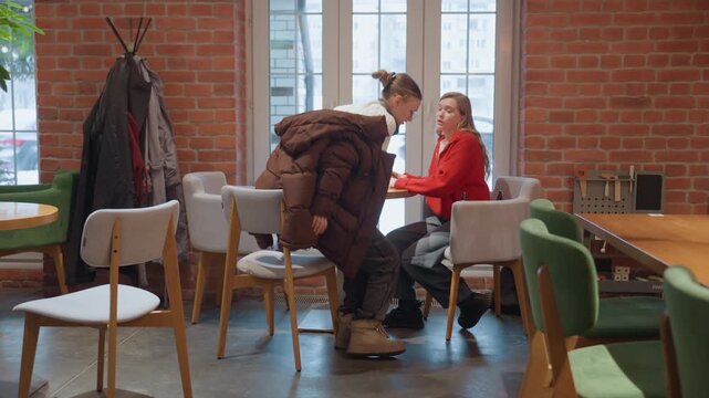 White woman waits alone at cafe. Red sweater sits near window with mug, looks thoughtful, friend arrives in brown puffer and joins at table, they hug and hold hands, warm brick interior, coat rack