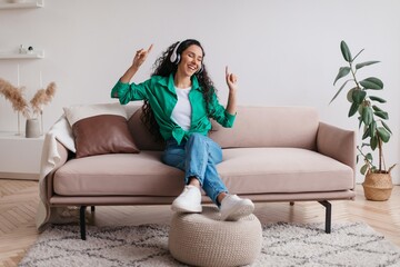 Obraz premium Smiling Young Woman Dancing And Enjoying Music While Sitting On Sofa In Cozy Living Room, Displaying Happiness And Joy In Indoor Comfort. Relaxed Home Lifestyle Concept.