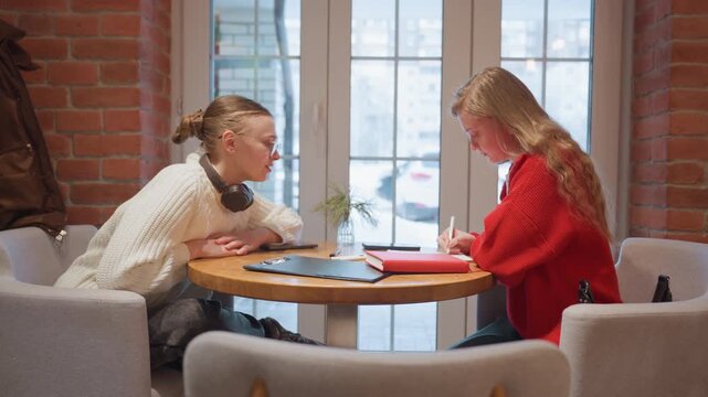 Caucasian women study in cozy cafe, focused tutor guiding student through notes at round table near brick window, headphones draped, red sweater, notebook, warm natural light, supportive atmosphere