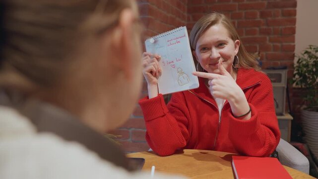 Young white woman in cozy cafe showing handwritten note to friend, wearing red sweater, smiling and revealing doodle portrait and short message, wooden table and brick backdrop, natural daytime light,