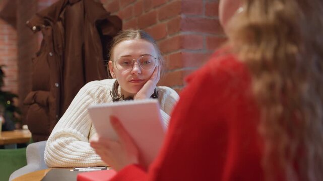White woman engaged in cafe meeting friendly interviewer in red sweater reads from tablet while listener reacts with thoughtful smile, exposed brick and window light, holiday decor adding warm tones,