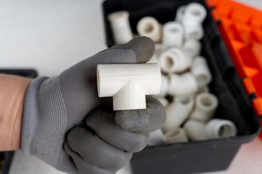 Hand in a grey protective glove holding a white polypropylene ppr tee fitting with an open toolbox blurred in the background.