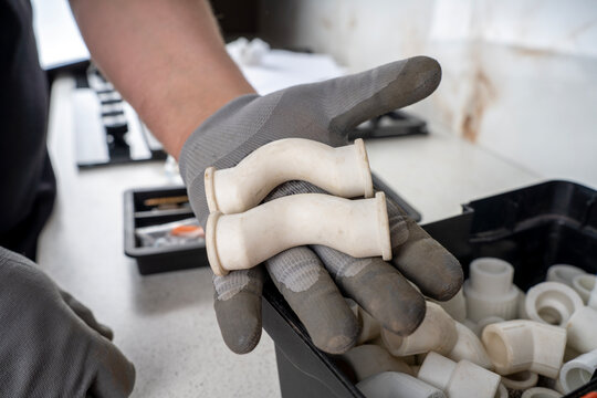Hand in a grey protective glove holding two white polypropylene ppr crossover pipe fittings over a plastic toolbox container.