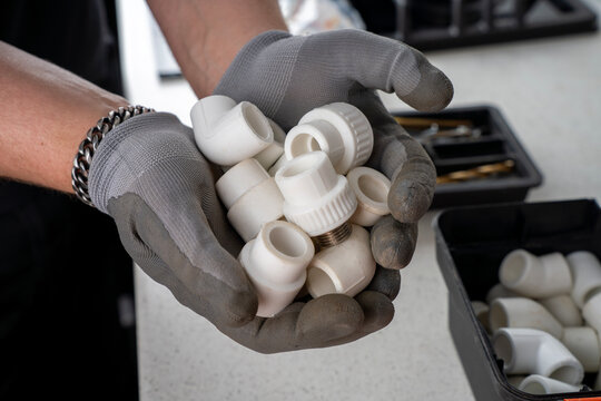Hands in grey protective work gloves holding a handful of white polypropylene ppr plumbing fittings, couplers, and elbows over a workshop table.