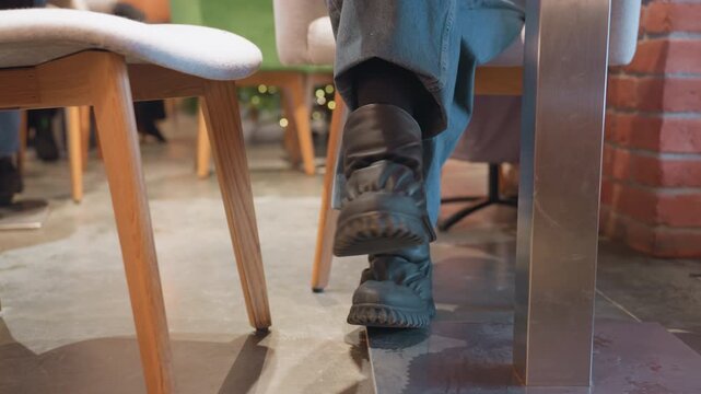 Cafe low angle boots resting under table freelancer legs crossed, black chunky shoes, denim pants, metal table base, wooden chairs, exposed brick wall, warm lighting, laptop on tabletop, calm leg