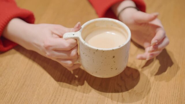 Hands cradle latte mug on cafe. Close shots of hands lifting and setting down mug on wooden table, red sweater sleeve, creamy latte surface and subtle steam, comforting morning ritual with relaxed