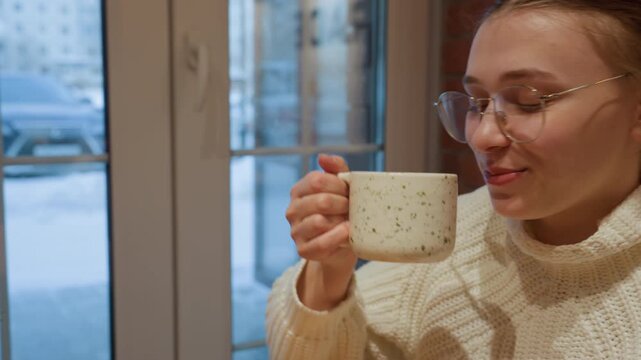 Cozy cafe conversation friends clinking speckled mugs. Caucasian woman in cream sweater clinks mug with companion on wooden table by snowy window, lifts warm ceramic cup, sips coffee with contented