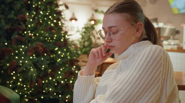 White woman in cozy cafe near Christmas tree, wearing chunky knit sweater and glasses, resting chin on hand while sipping latte, warm bokeh lights, calm reflective mood, indoor holiday interior