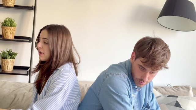 A young couple sits back-to-back on a sofa, engaged in a heated argument. They appear upset and frustrated, showcasing a relationship problem or disagreement at home due to a lack of communication.