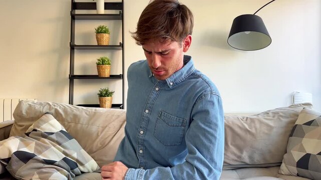 A young man experiences back and neck pain while sitting on the sofa at home, stretching to find relief from the discomfort. He attempts to crack his back and neck to alleviate tension and aches.