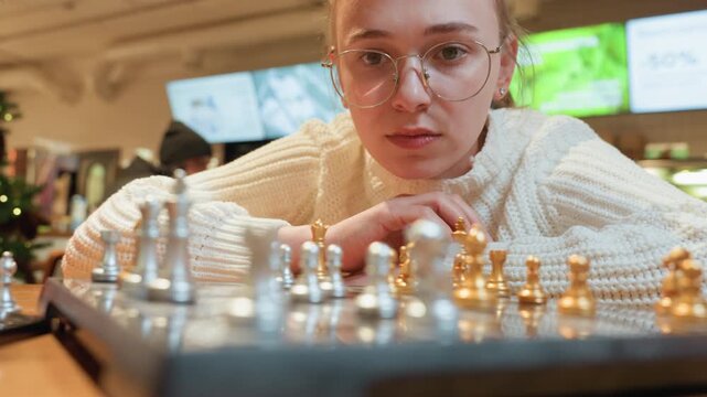 Caucasian woman plays chess match at cafe table, focused gaze through glasses, rows of gold and silver pieces in foreground, deliberate hand reaches for tactical move, barista and patrons blurred