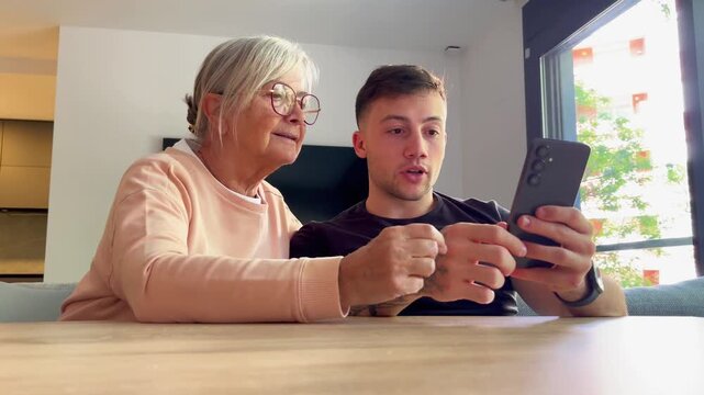 A young man patiently teaches an elderly woman how to use a smartphone, bridging the generational gap through technology and sharing a moment of connection and learning at home.