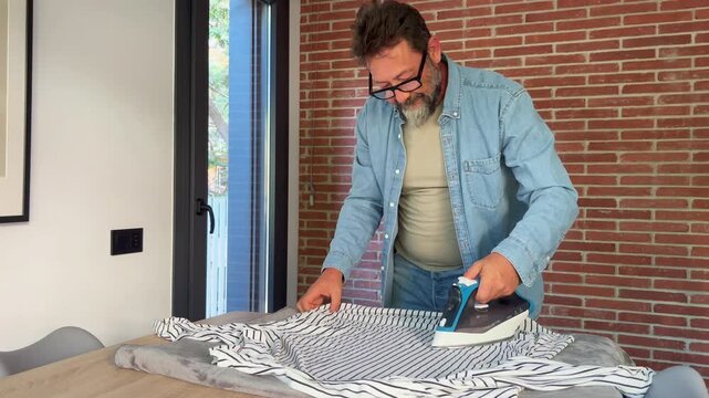 A man meticulously irons a striped shirt on a wooden table, performing a common household chore. The focus is on the routine task of keeping clothes neat and tidy at home.