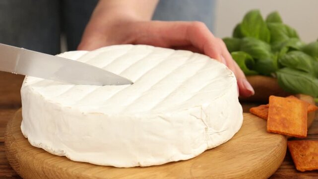 Woman cutting delicious brie cheese and showing piece at wooden table against grey background, closeup