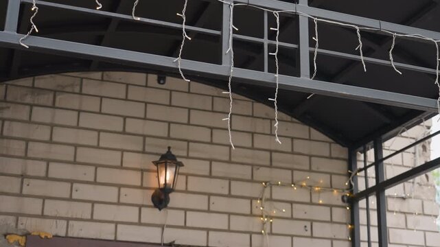 Urban brick facade with metal canopy and glowing lantern under hanging string lights, industrial steel frame against neutral beige masonry, overcast sky, quiet city atmosphere, cozy ambient glow