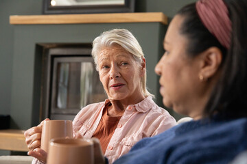 Obraz premium Two mature women sitting on couch in living room holding tan mugs, one wearing pink headband