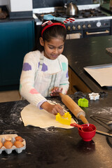 Indian school-age girl pressing yellow cookie cutter into rolled dough in kitchen wearing apron © wavebreak3