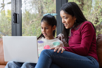 Indian mother leaning over school-aged daughter, helping with white laptop on leather sofa at home © wavebreak3