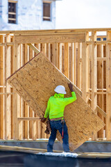 Fototapeta premium A vertical view from behind a construction worker wearing a hard hat and a high-visibility green shirt while carrying a large sheet of OSB plywood paneling on a building framing site, USA