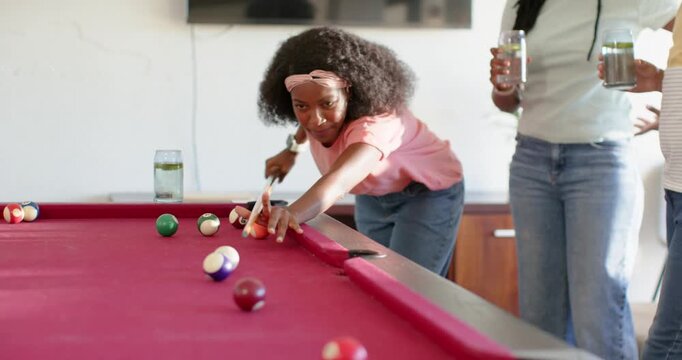 African American friends cheering pink-shirt woman aiming and hitting cue on red pool table at home