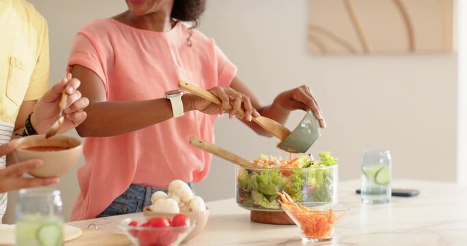African family woman in coral-top pouring dressing, tossing salad with tongs in kitchen for meal