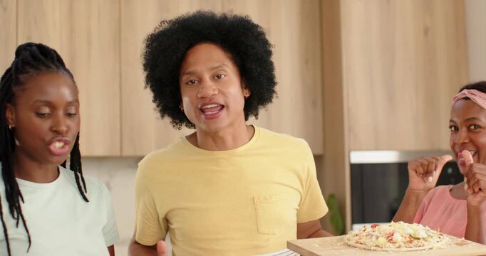 African American trio in kitchen, man in yellow holding board, guiding pizza making with vegetables
