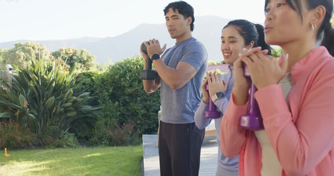 Diverse friends performing syncing squats on deck, holding dumbbells and kettlebell for workout