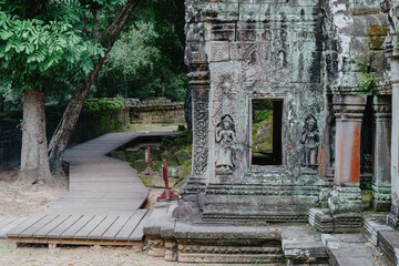 Fototapeta premium Cambodia, Siem Reap. A wooden walkway leads through the ancient ruins of Ta Prohm temple, overgrown with moss and jungle vegetation.