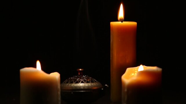 Three magic Christmas wax candle flames calm burning in home in white smoke from incense aroma stick on black dark background with copy space. Closeup yoga, spa, wellness, relaxation. No people