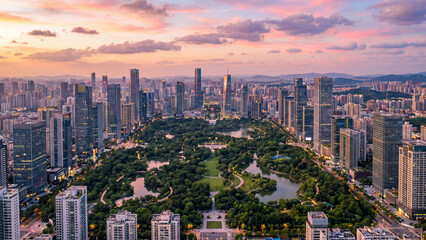 Fototapeta premium Metropolitan park glows under a vibrant pink and purple sunset sky while surrounded by a modern skyline of high-rise glass buildings and dense urban infrastructure in a bustling financial district.
