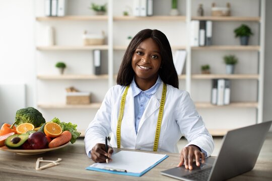 Smiling Black Female Nutritionist Working At Office, Taking Notes While Surrounded By Fresh Fruits And Vegetables. Promoting Healthy Lifestyle And Nutritional Guidance. Weight Loss Concept.