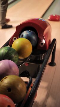 Vertical close-up video of colorful bowling balls on a ball return rack in a bowling alley, shallow focus with anonymous player legs in background and indoor leisure game atmosphere