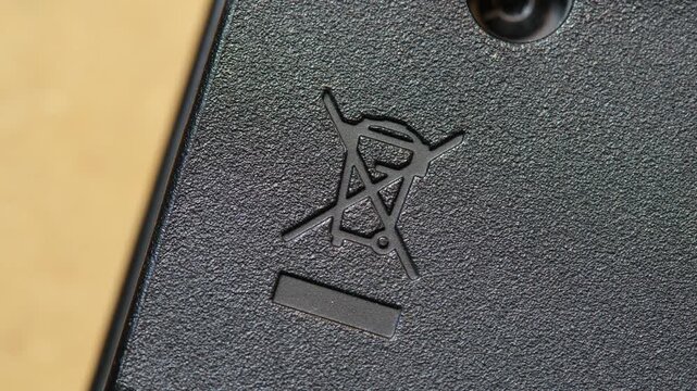 Slow macro pan across crossed-out wheeled bin WEEE recycling symbol on black textured electronic device casing, close-up of disposal warning mark, blank label, screw and e-waste compliance detail