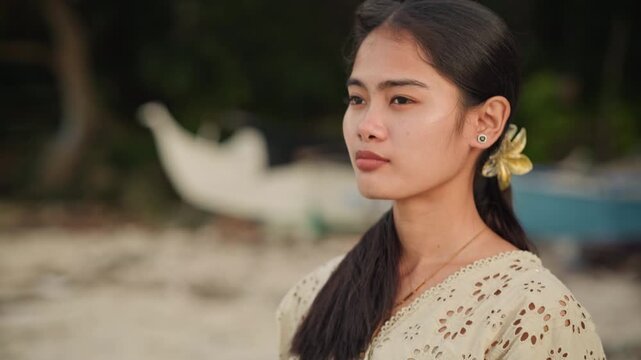 Young woman on beach looking outward, adjusts earring and tucks hair, yellow flower behind ear near small boat and palm silhouettes, soft golden hour light, hopeful confident expression and calm