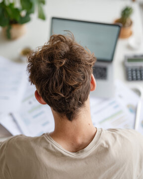 Young man works at home office desk. Curly brown hair frames his focused back view. Laptop sits beside scattered charts and documents. Natural light brightens the calm, organized workspace