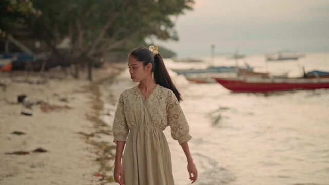 Asian girl walking along beach shore, pausing to scan horizon, carrying printed tote and striped towel, picking up towel near moored boats, turning toward village path, warm dusk light, reflective yet